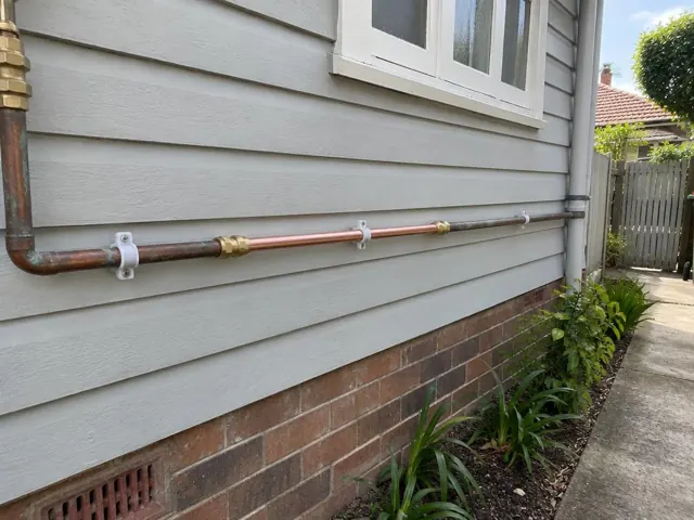 Repaired copper water pipe along a weatherboard house exterior wall with brick base in Belfield
